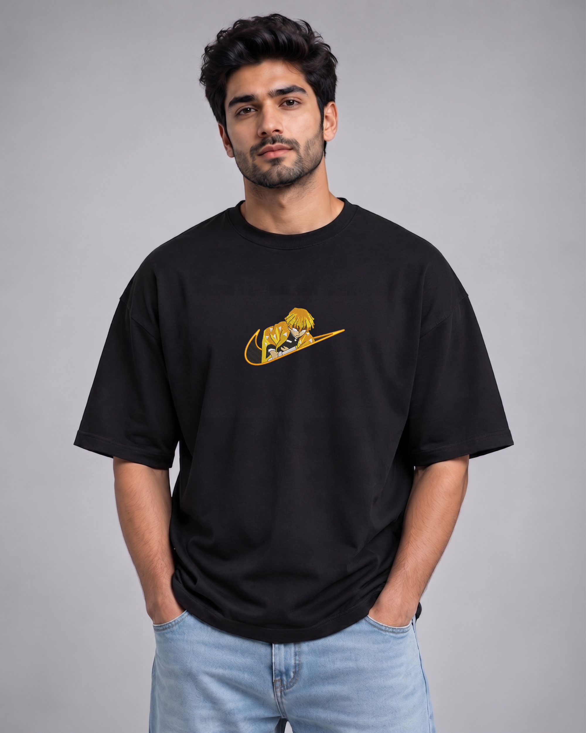 A man wearing a black oversized t-shirt with a yellow graphic on the chest, paired with light blue jeans, standing against a solid gray studio background.
